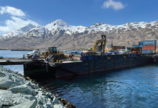 Barge on water loaded with cargo including heavy equipment. Snow capped mountains in background.