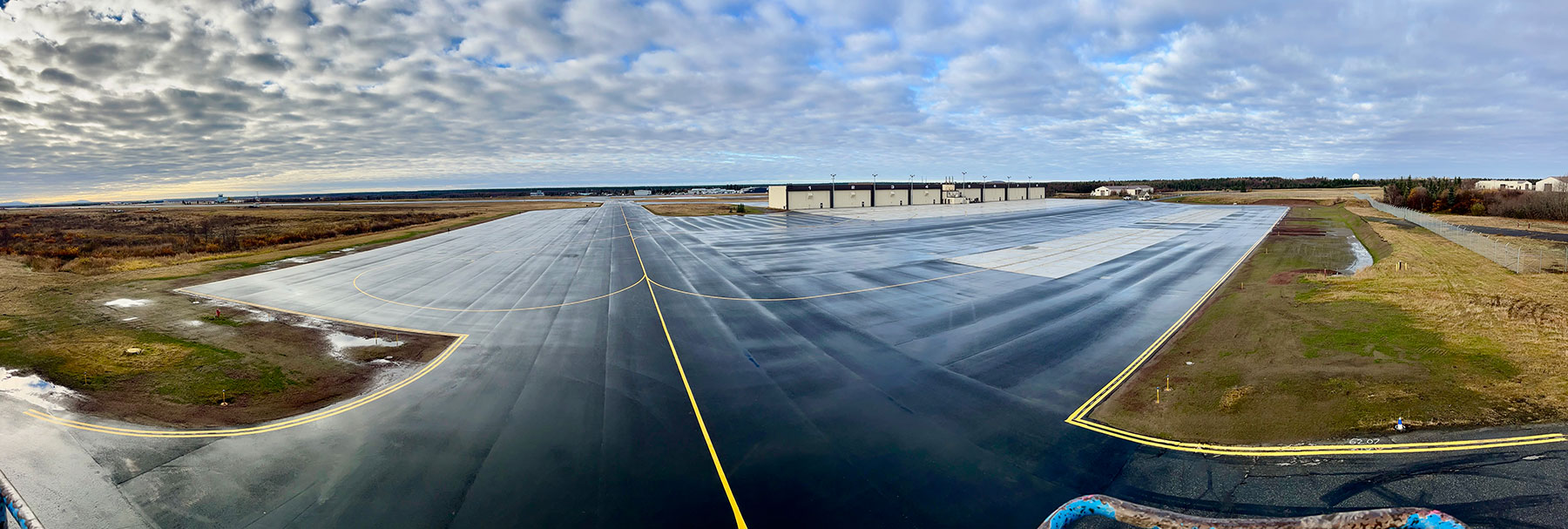 Panoramic view of newly repaved taxiway and apron at King Salmon Airport in King Salmon, Alaska, with fresh yellow markings leading to a row of aircraft hangars under a cloudy sky.